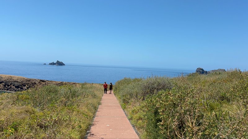 Two people walking along a paved path through scrubby vegetation toward the sea, with small rocky islets visible offshore
