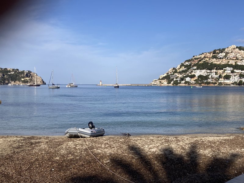 View across the anchorage at Port d'Andratx: a few sailboats at anchor, a small red lighthouse on the breakwater, and a hillside covered in white villas, with an inflatable dinghy beached in the foreground