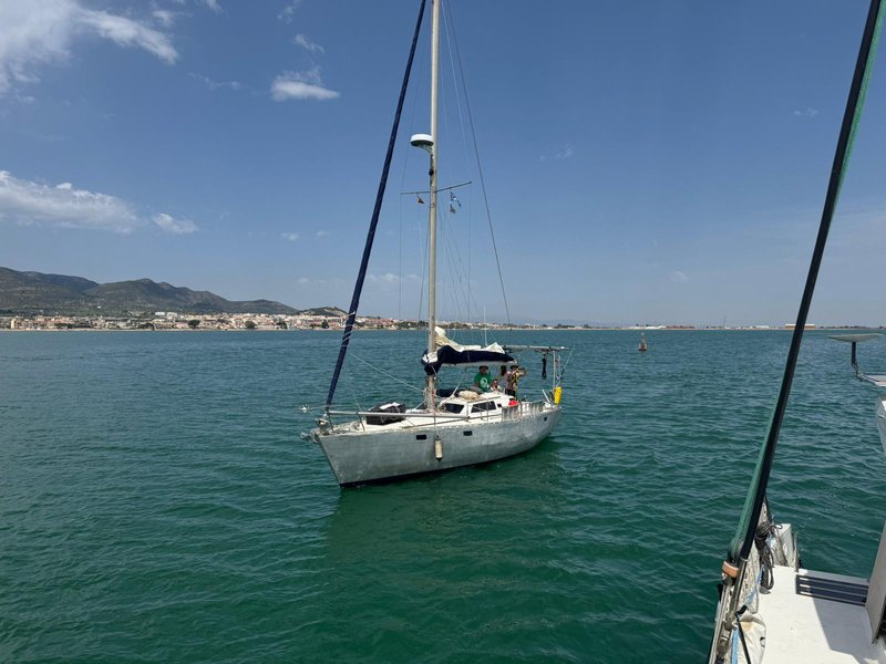 Tinarasia motoring away under bare poles across flat green-blue water, a low coastal town and hills in the background, seen from another boat