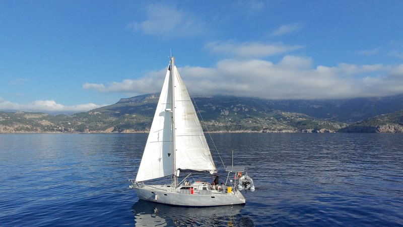 Aerial view of Tinarasia under sail, mainsail and headsail up, approaching a mountainous coastline under a partly cloudy sky