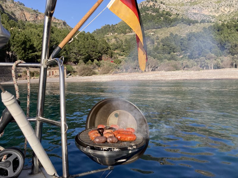 A hanging BBQ grill off the stern rail, sausages and mushrooms cooking over the coals with smoke rising, a German flag flying above and a pine-covered cove with a pebble beach behind