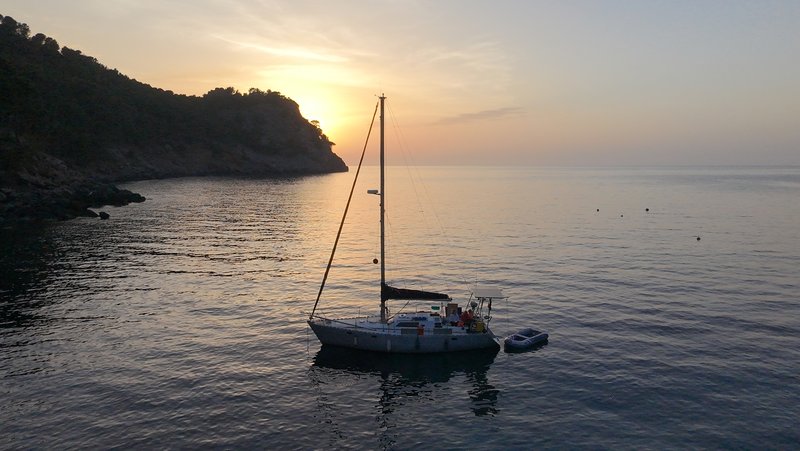 Tinarasia at anchor in a calm cove at sunset, with a dinghy alongside and a headland silhouetted against the glowing sky