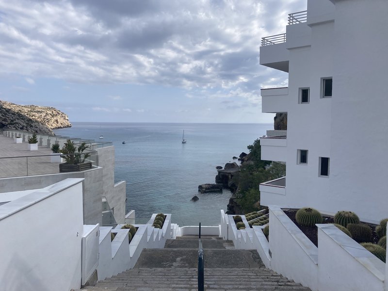 View down a white staircase past modern villas to a small rocky cove with a sailboat anchored offshore