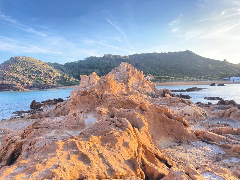 Jagged reddish-orange rock formation rising from turquoise water, with green hills and a white house in the background at low sun