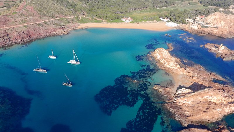 Aerial view of four sailboats anchored in a turquoise bay with a sandy beach, backed by rocky reddish cliffs and green hills