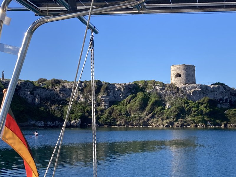 View from the boat's stern rail across the water to a round stone watchtower on a green, rocky headland, with a German flag flying in the foreground