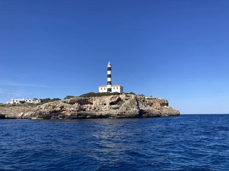 A black-and-white striped lighthouse atop a rocky headland, seen from the water under a clear blue sky