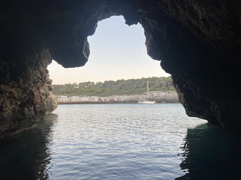 View out through the mouth of a dark sea cave toward a sailboat anchored in a sunlit bay lined with pine trees