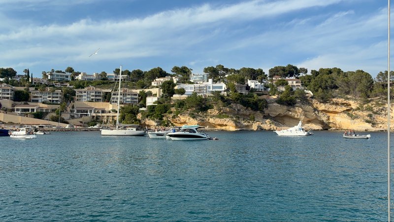 Several boats anchored in a small bay backed by sandy cliffs and white villas among pine trees