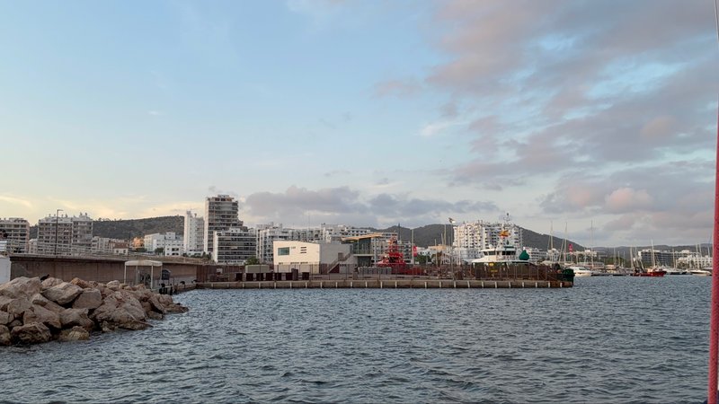A harbor breakwater with a pile of rocks in the foreground and a dense skyline of white apartment buildings behind, under a pastel evening sky