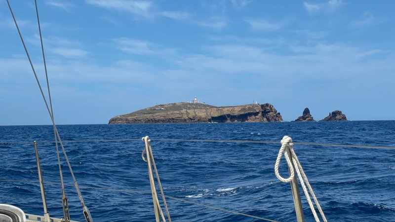 A rocky island with a lighthouse, seen across open water past the boat's bow rail and rigging