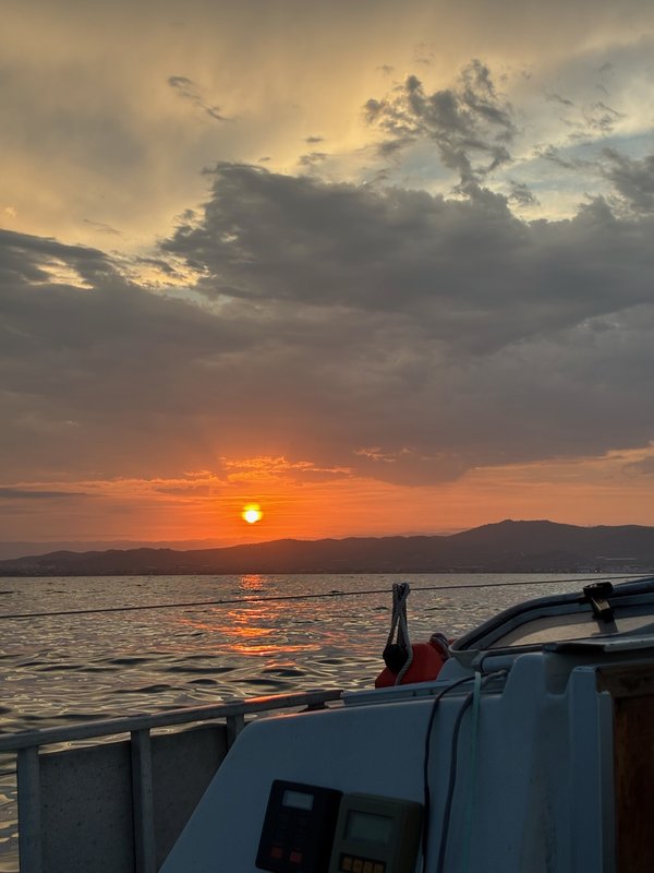 A glowing orange sunset over a mountainous coastline, seen from the boat's cockpit with instruments and railing in the foreground