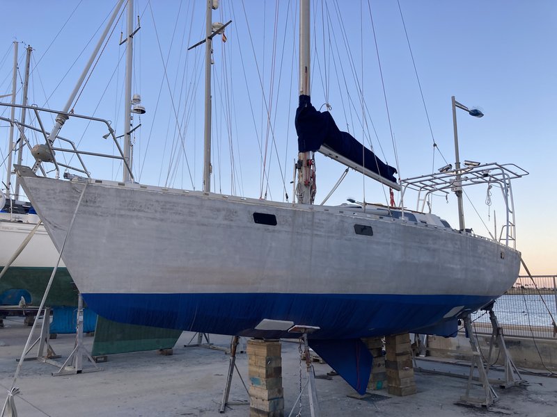 Tinarasia hauled out on stands in the boatyard, her hull sanded bare with fresh blue antifouling on the bottom, masts and rigging silhouetted against a dusky sky